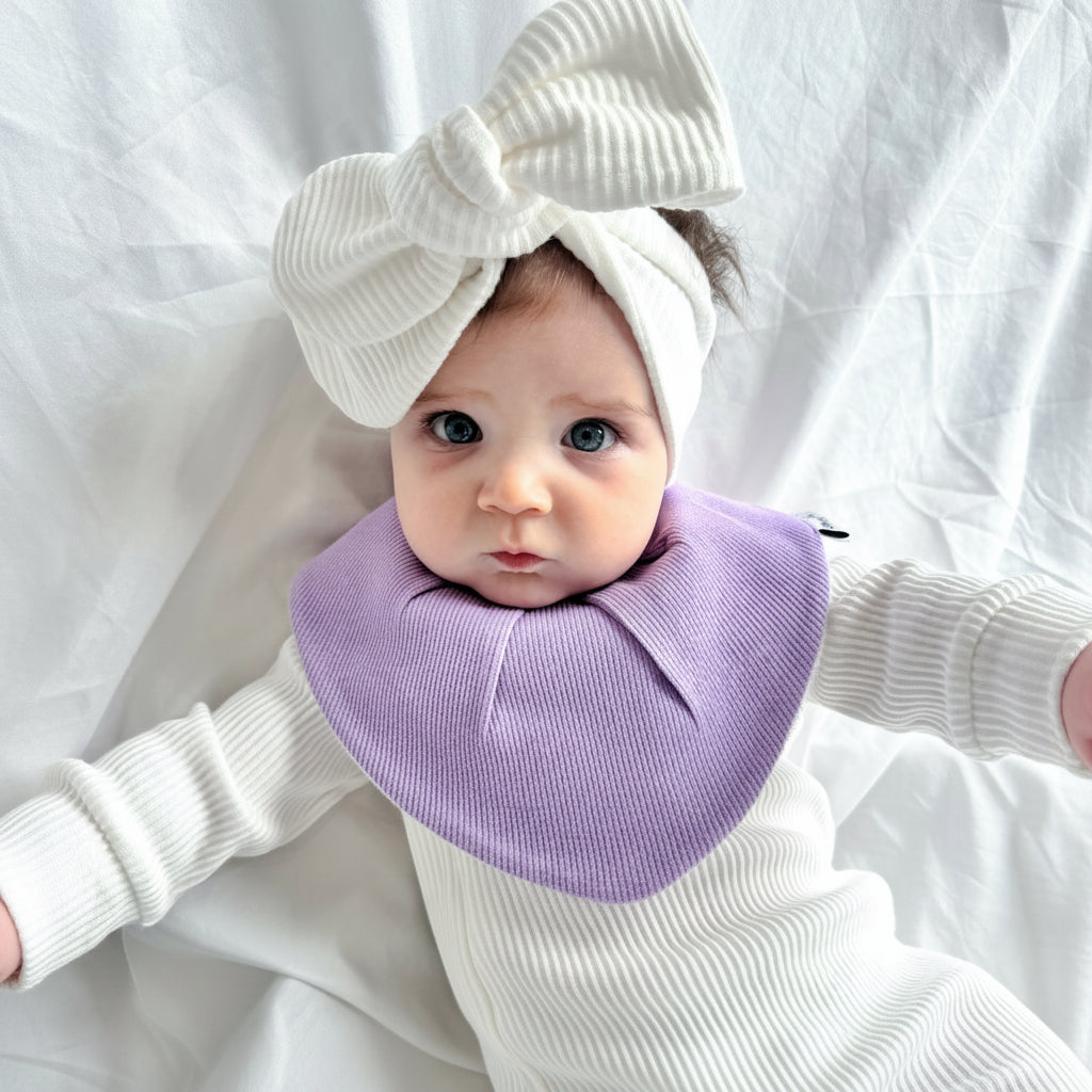 A baby wearing a white ribbed long-sleeve outfit, a large white bow headband, and a lavender lilac bib, lying on a white fabric background.
