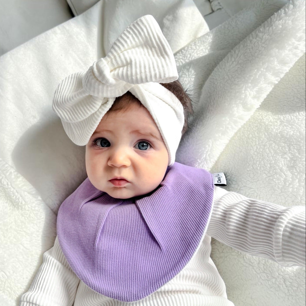 A baby lying on white bedding, wearing a white ribbed long-sleeve bodysuit, a large white bow headband, and a lavender purple bib.
