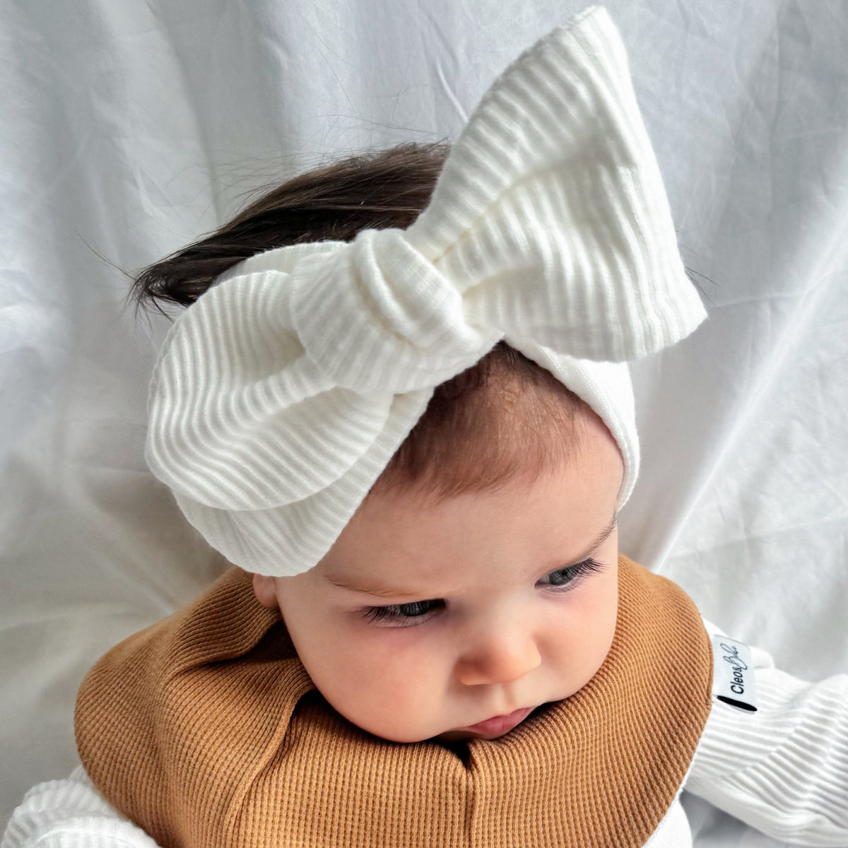 A baby wearing a white ribbed long-sleeve footed romper, a tan waffle-knit bib, and a large white ribbed bow headband, lying on a white fabric backdrop.