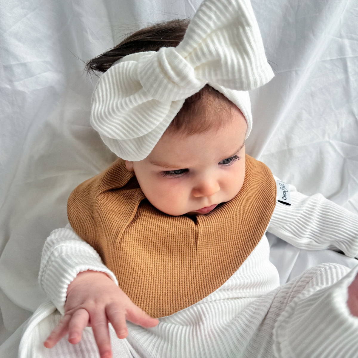 A baby wearing a white ribbed long-sleeve footed romper, a tan waffle-knit bib, and a large white ribbed bow headband, lying on a white fabric backdrop.