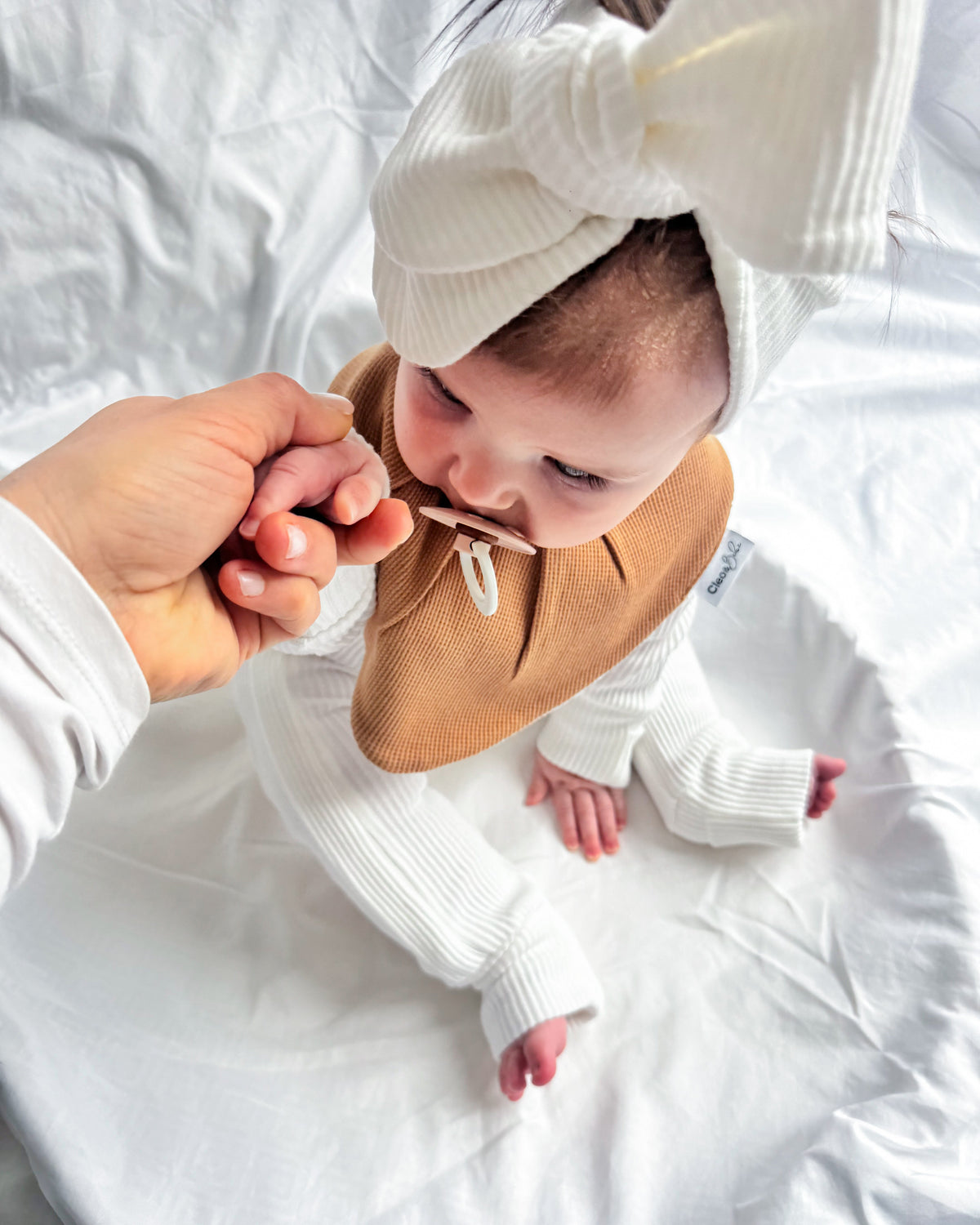 A baby wearing a white ribbed long-sleeve bodysuit, a tan bib, and a white knotted headband sits on white bedding while an adult hand gently holds the baby's hand.