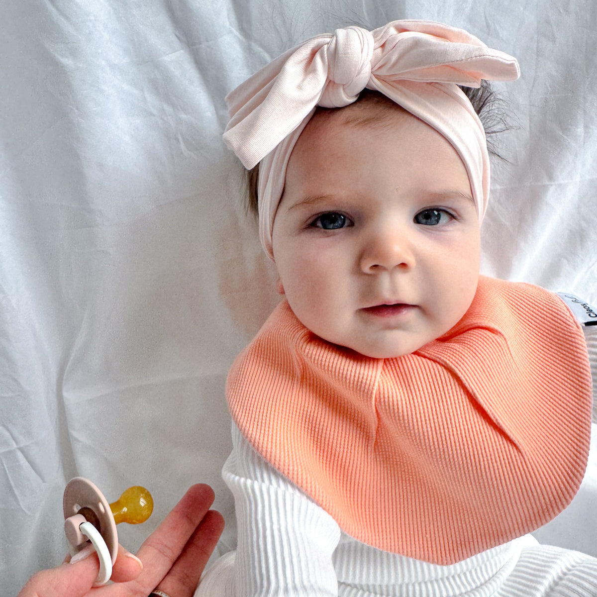 A baby wearing a peach ribbed bib and a pale pink knotted headband, lying on a white fabric surface. A hand holding a pacifier is visible at the lower left.