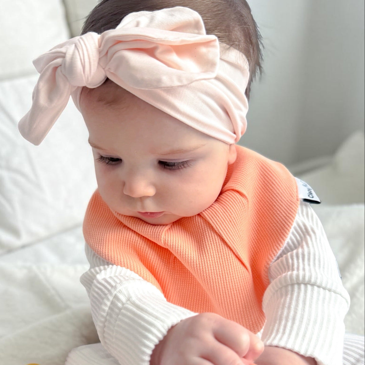 A baby wearing a soft peach-pink knotted headband, a ribbed peach bib, and a white long-sleeve top sits on a white blanket while an adult hand offers a pacifier.