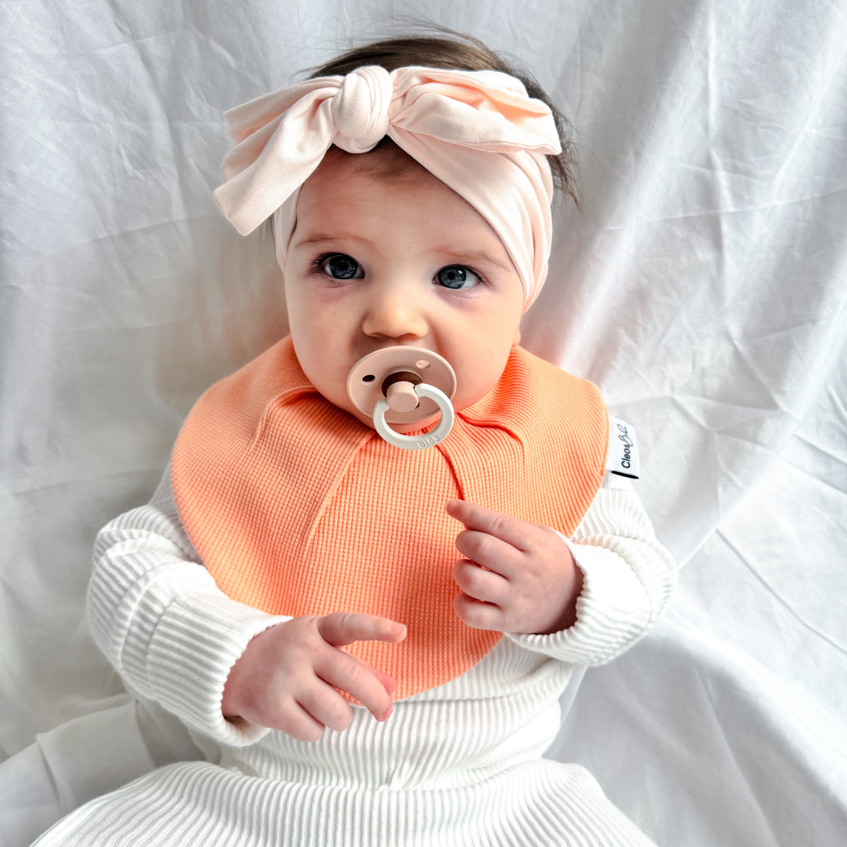 A baby wearing a white ribbed one-piece outfit, a peach waterproof bib, and a matching peach-orange bow headband, sitting against a white fabric backdrop.