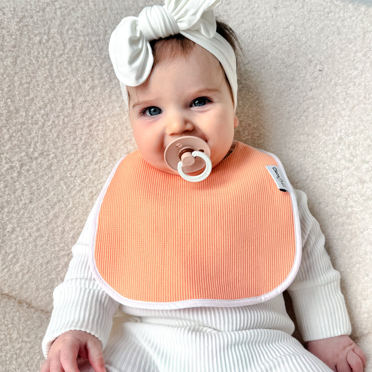 A baby wearing a white ribbed one-piece outfit, a white bow headband, and a peach-colored bib, sitting on a cream-colored textured chair.