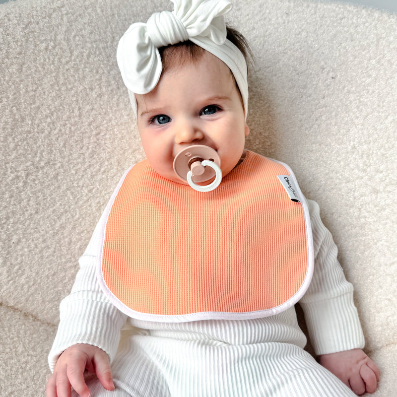A baby wearing a white ribbed one-piece outfit, a white bow headband, and a peach-colored bib, sitting on a cream-colored textured chair.