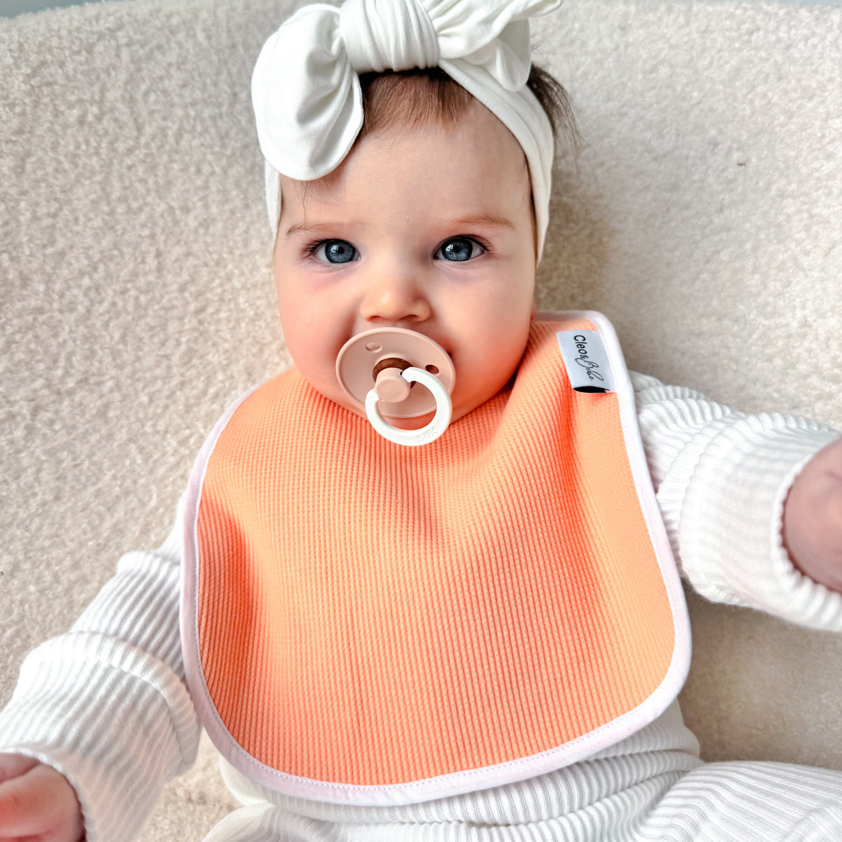 A baby wearing a white ribbed bodysuit, a white knotted bow headband, and a peach waffle waterproof bib, lying on a beige textured couch.