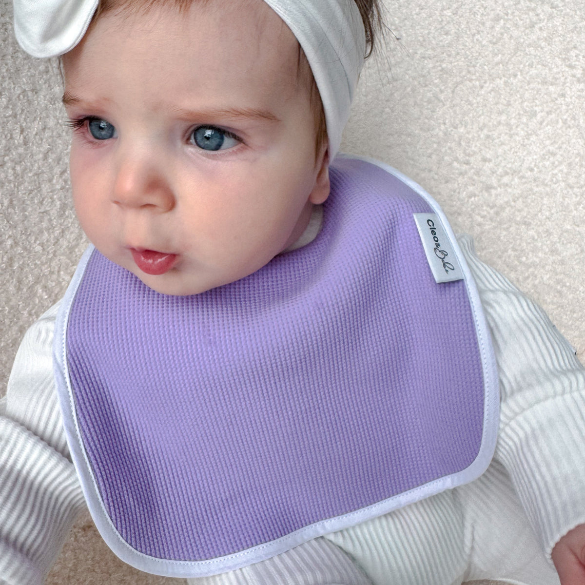 A baby wearing a lilac ribbed bib with a white border and a matching white bow headband, lying on a beige textured blanket.