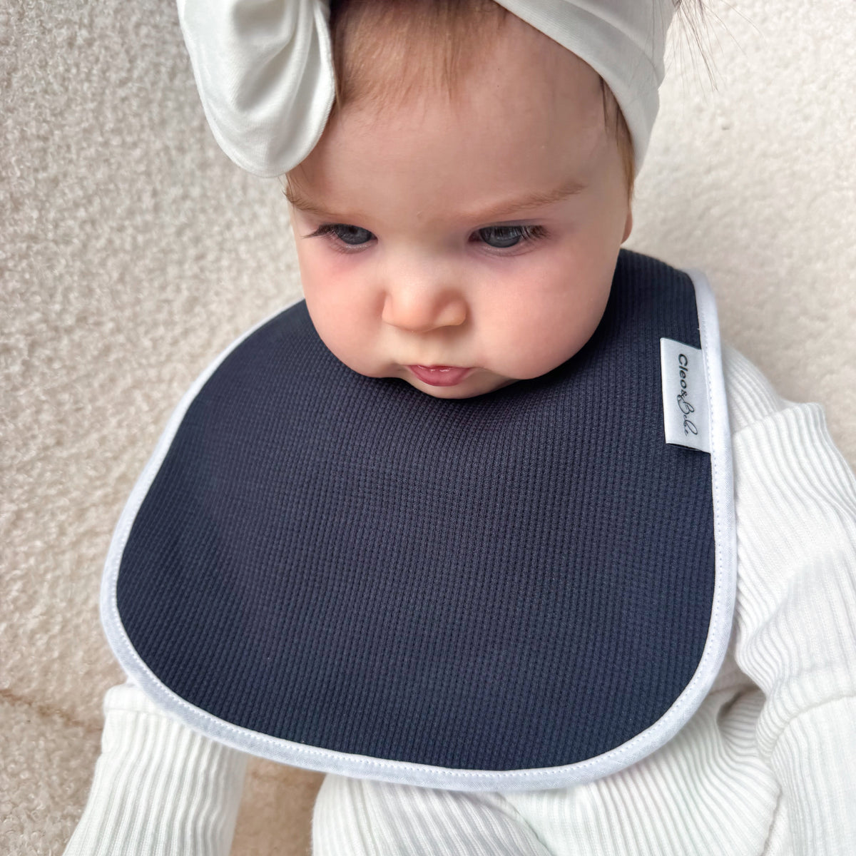 A baby wearing a white ribbed outfit and a matching white bow headband, sitting on a cream textured surface and wearing a dark navy bib with a white border. The bib has a small white brand tag on the right side that reads 'Bebamour'.