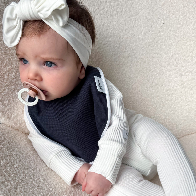 A baby wearing a ribbed cream footed one-piece with a matching bow headband and a navy bib, lying on a textured cream chair.