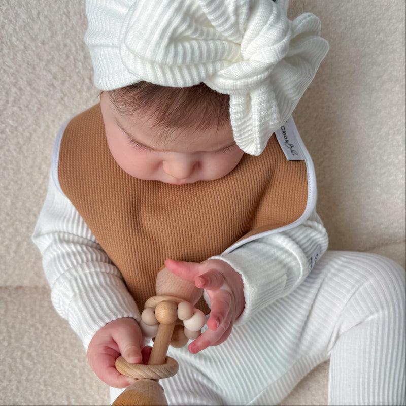 A baby wearing a white ribbed footed one-piece, a tan bib, and a large white bow headband sits on a textured cream surface while holding a wooden teether.