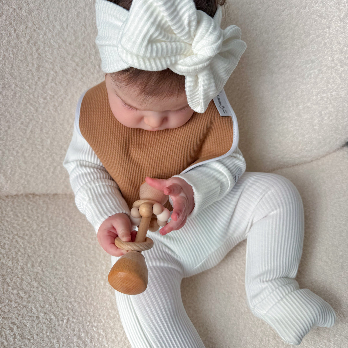 A baby wearing a white ribbed footed one-piece, a tan bib, and a large white bow headband sits on a textured cream surface while holding a wooden teether.
