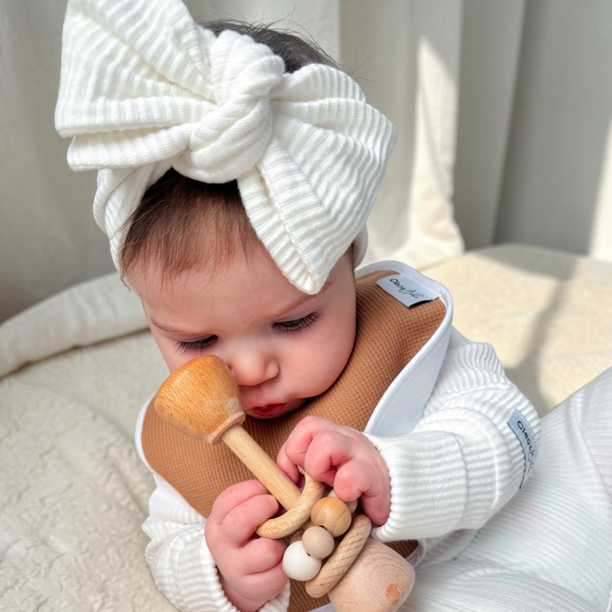 A baby wearing a white ribbed long-sleeve outfit and a large white bow headband, lying on a cream textured blanket and holding a wooden mushroom teether.