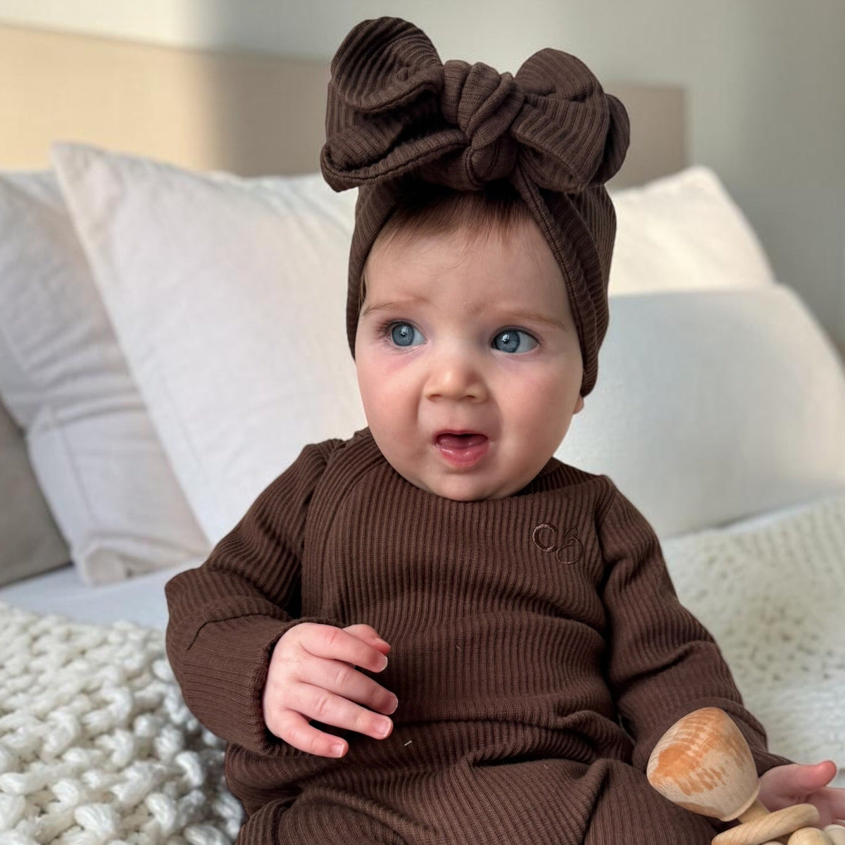 A baby wearing a matching brown ribbed one-piece outfit and large bow headband, sitting on a bed with white bedding and holding a wooden teether.
