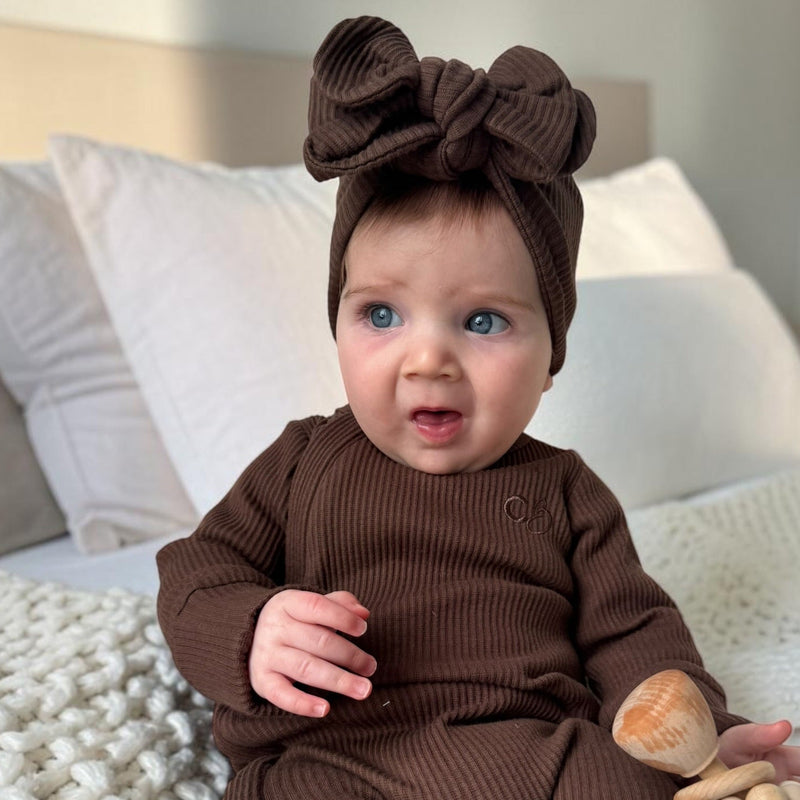 A baby wearing a matching brown ribbed one-piece outfit and large bow headband, sitting on a bed with white bedding and holding a wooden teether.
