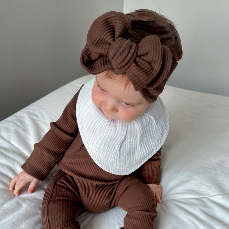 A baby wearing a brown ribbed long-sleeve outfit with matching pants and a large matching headband, plus a white ribbed bib, sitting on a white bed.