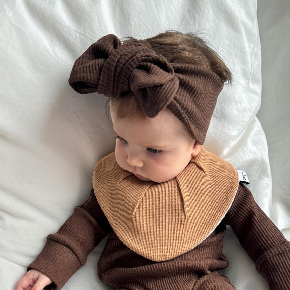 A baby lying on white bedding wearing a ribbed brown one-piece outfit with a large tan bib and matching brown bow headband.