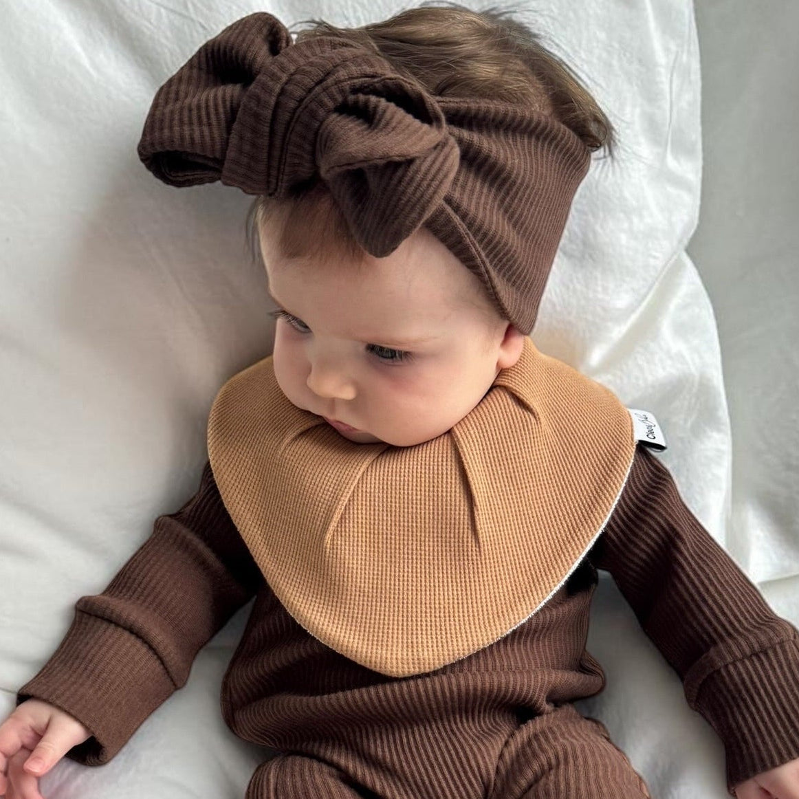 A baby lying on white bedding wearing a ribbed brown one-piece outfit with a large tan bib and matching brown bow headband.