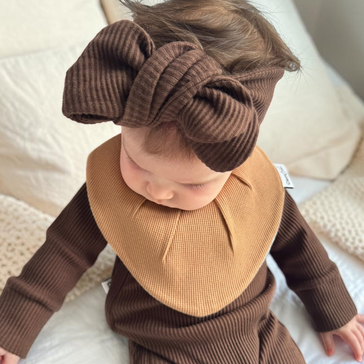 A baby wearing a brown ribbed long-sleeve one-piece outfit, a tan ribbed bib, and a matching brown ribbed bow headband, sitting on a white bed.