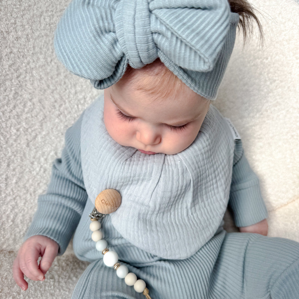 A baby wearing a light blue ribbed one-piece outfit with a matching bow headband and bib, with a wooden bead pacifier clip, lying on a soft cream-colored chair.
