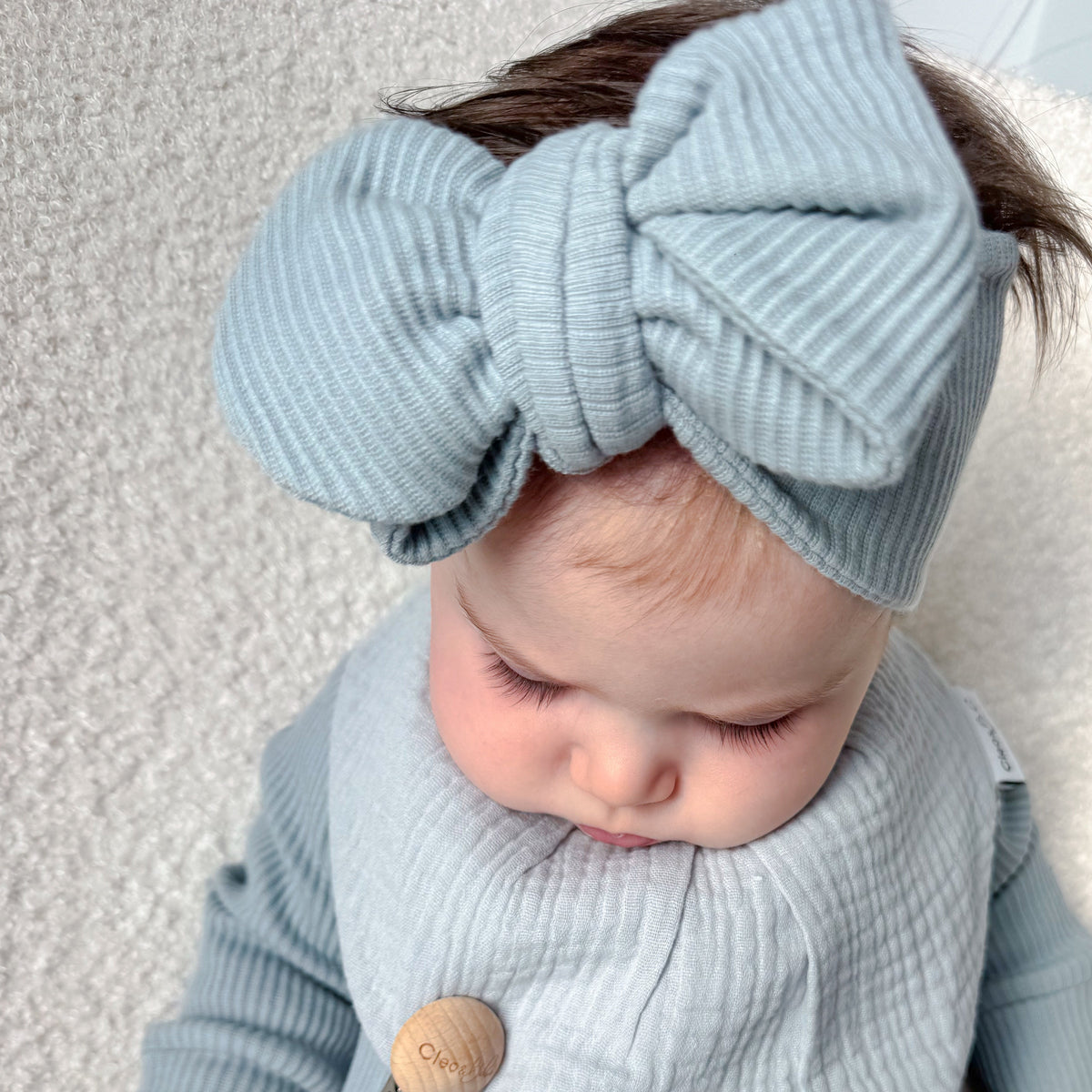 A baby wearing a light blue ribbed one-piece outfit with a matching bow headband and a wooden bead pacifier clip, lying on a soft cream-colored blanket.