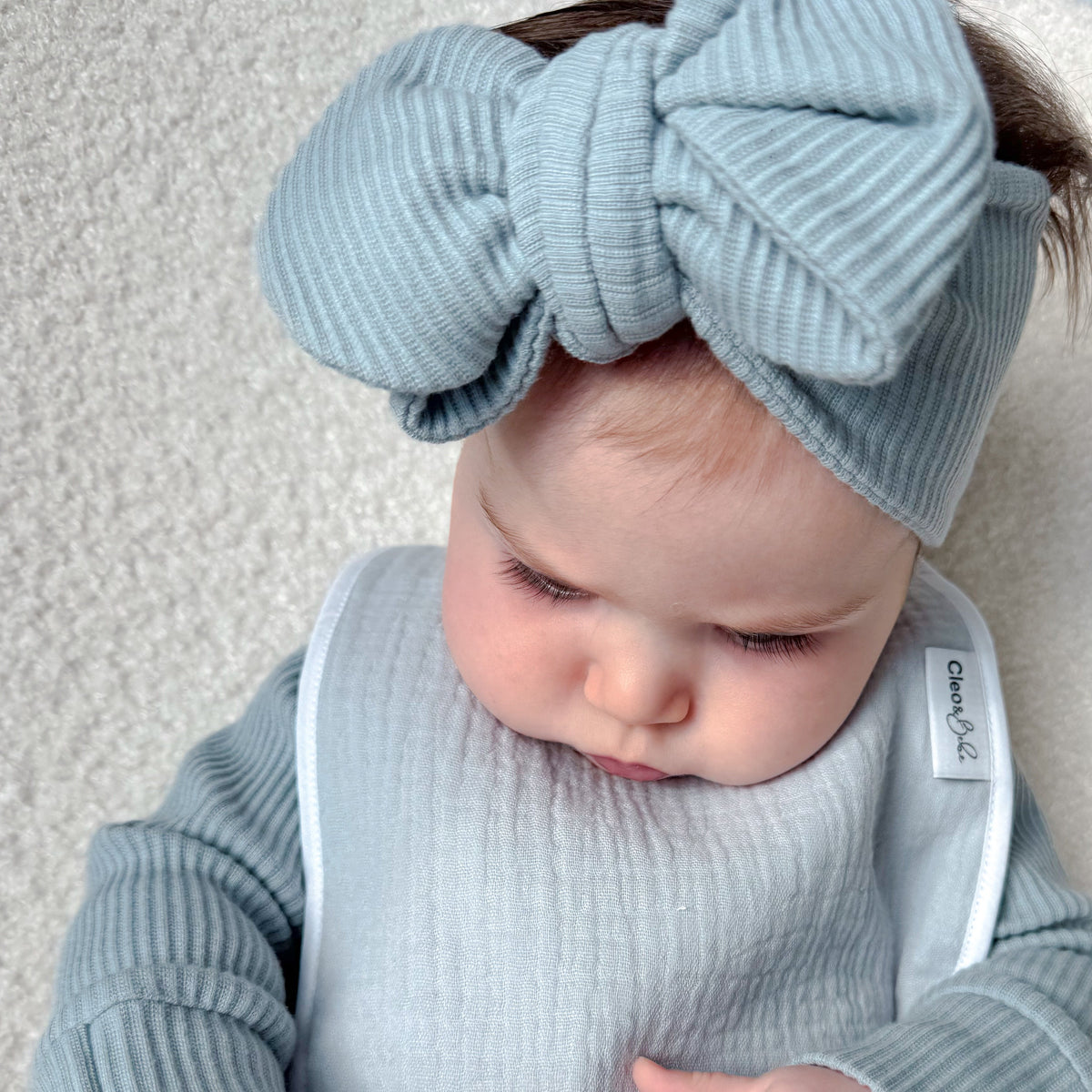A baby wearing a light blue ribbed long-sleeve outfit, matching bow headband, and bib, holding a pacifier clip toy on a cream textured blanket.