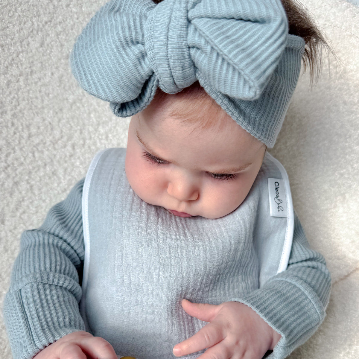A baby wearing a light blue ribbed long-sleeve outfit, matching bow headband, and bib, holding a pacifier clip toy on a cream textured blanket.