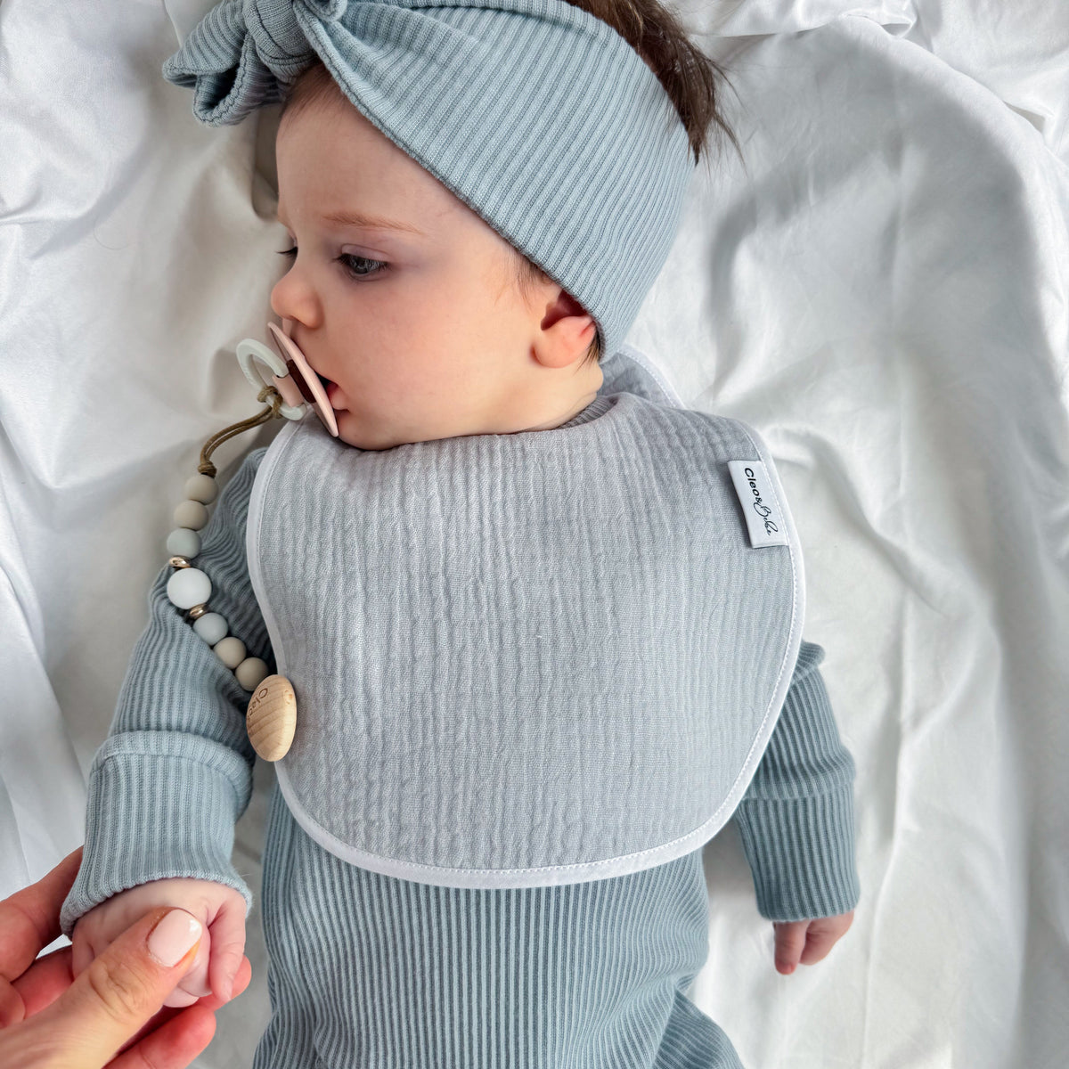 A baby lying on white bedding wearing a light blue ribbed long-sleeve bodysuit, matching bow headband, and bib. A pacifier clip with wooden beads is attached to the bib, and an adult hand is holding the baby's hand.