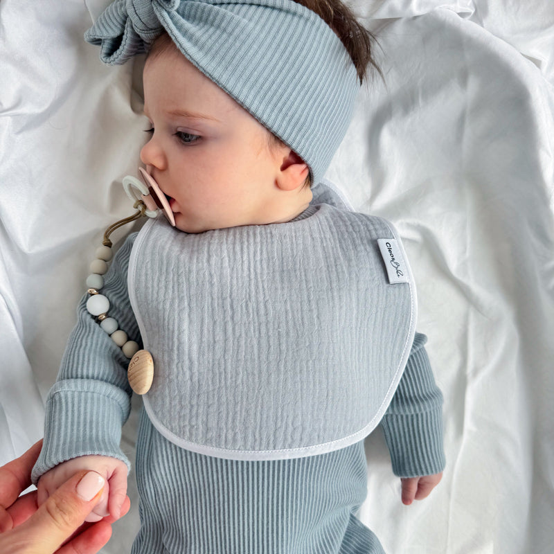 A baby lying on white bedding wearing a light blue ribbed long-sleeve bodysuit, matching bow headband, and bib. A pacifier clip with wooden beads is attached to the bib, and an adult hand is holding the baby's hand.