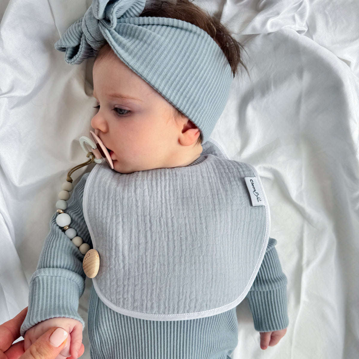 A baby lying on white bedding wearing a light blue ribbed long-sleeve bodysuit, matching bow headband, and bib. A pacifier clip with wooden beads is attached to the bib, and an adult hand is holding the baby's hand.