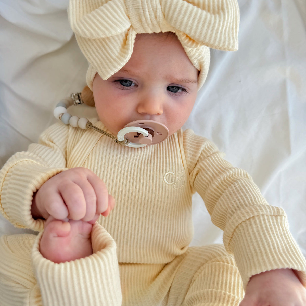A baby wearing a cream ribbed one-piece jumpsuit and matching large bow headband, lying on a white fabric background.