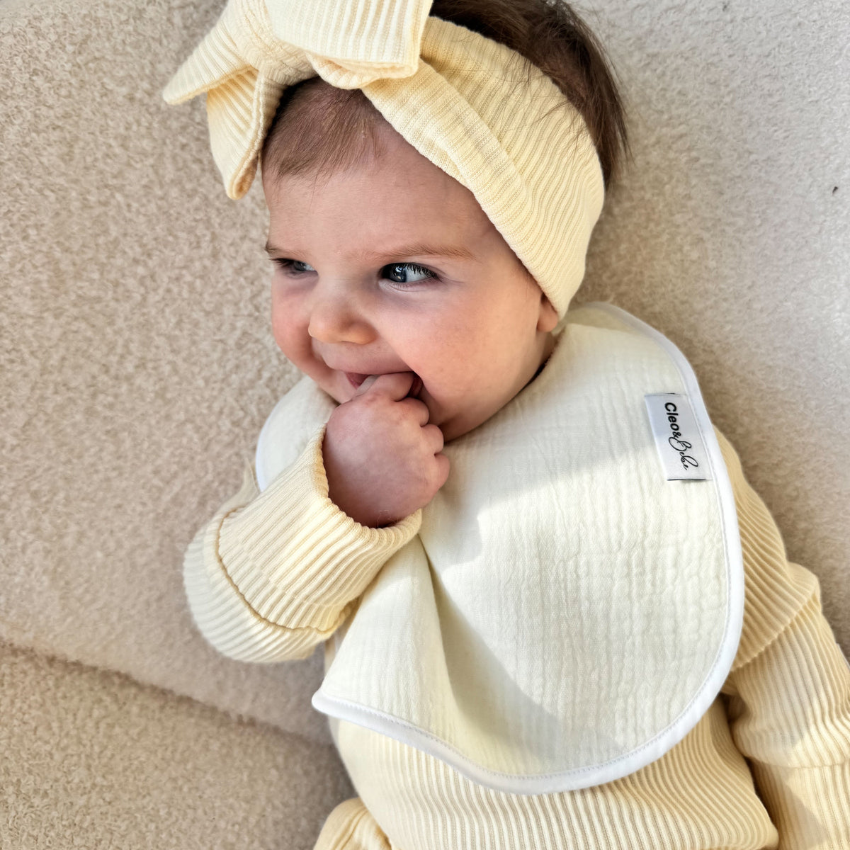 A baby wearing a lemon-colored ribbed outfit, matching bow headband, and bib, sitting on a textured beige sofa.