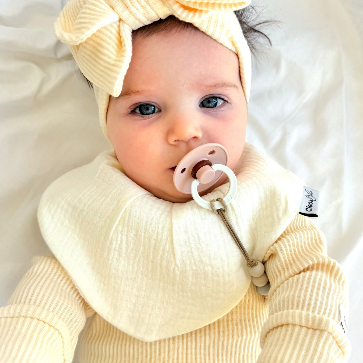 A baby lying on white bedding, wearing a pale yellow ribbed outfit with a large matching bow headband and a wide bib. The baby has a pacifier in their mouth.
