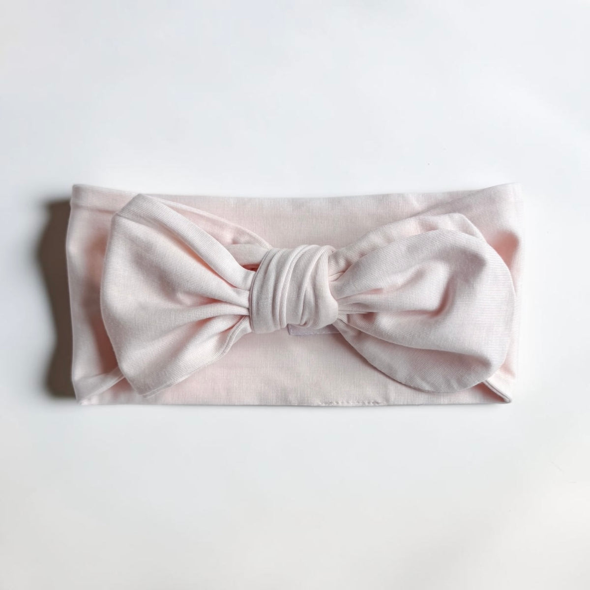 A pale pink fabric headband with a large bow in the center, laid flat on a white background.