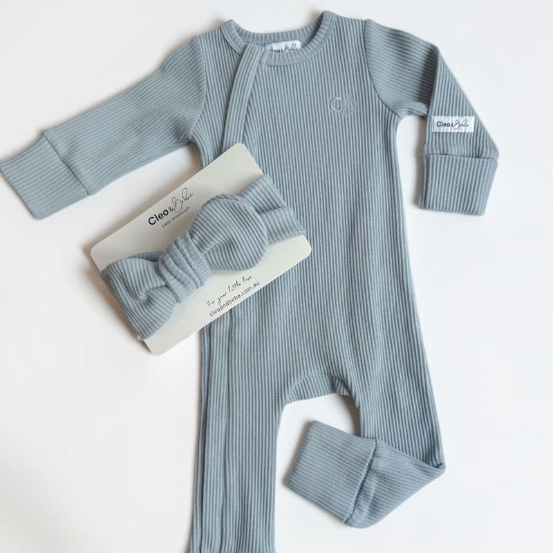 A baby's ribbed long-sleeve footed romper in dusty blue-gray, shown with a matching bow headband and a small branded tag on a white background.