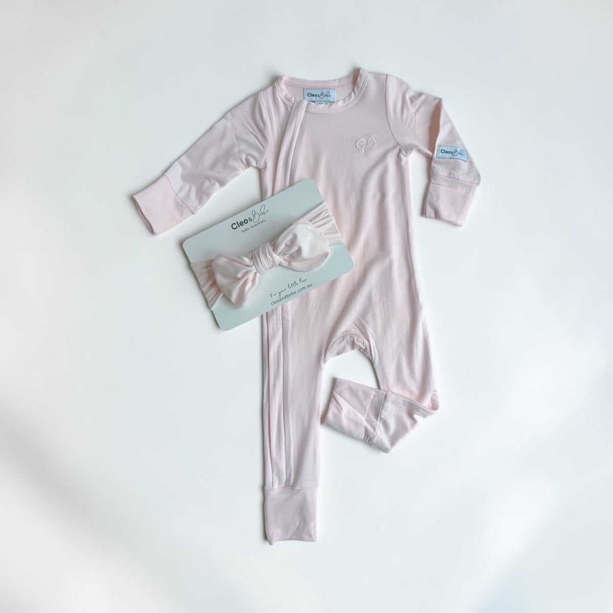 A baby's long-sleeve footless onesie in light pink, laid flat on a white background next to a matching pink bow headband on its packaging card. The brand 'Cleo & bebe' is visible on the packaging.