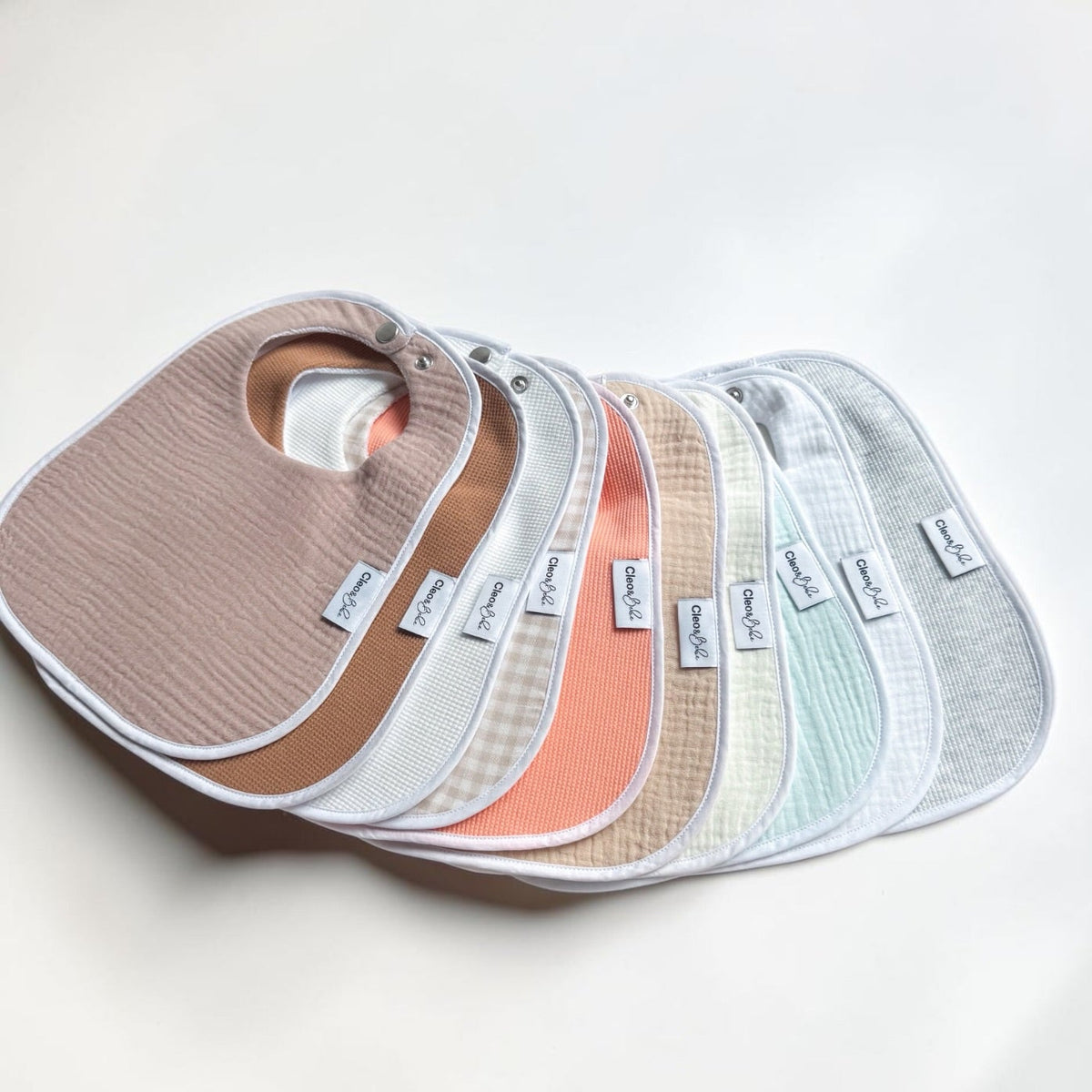 10 reusable muslin baby bibs in soft neutral and pastel colors, fanned out on a light off-white background. The brands 'Bib & Co' are visible on the small white labels attached to each bib.