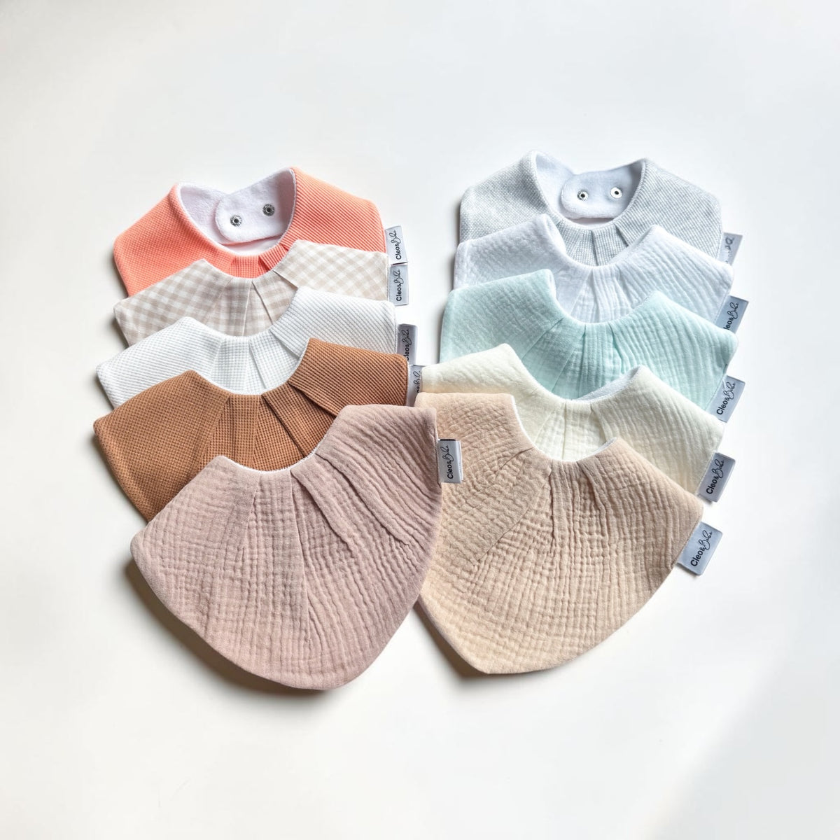Ten soft baby bibs in pastel colors arranged in two overlapping groups on a light neutral background. The bibs include coral, beige-and-white check, white, mint, cream, and blush pink, with a ribbed waffle texture and small fabric tags visible on the sides.